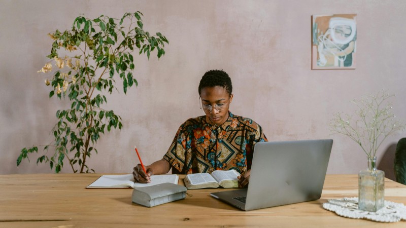 A person reading a book and taking notes at a wooden desk with a laptop indoors., tags: für - pexel