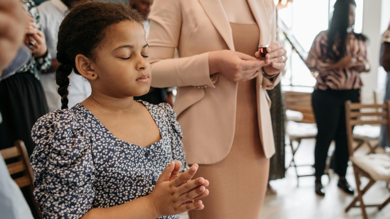 A young girl in a floral dress silently prays during a community gathering indoors., tags: und glaubensgemeinschaften - pexel